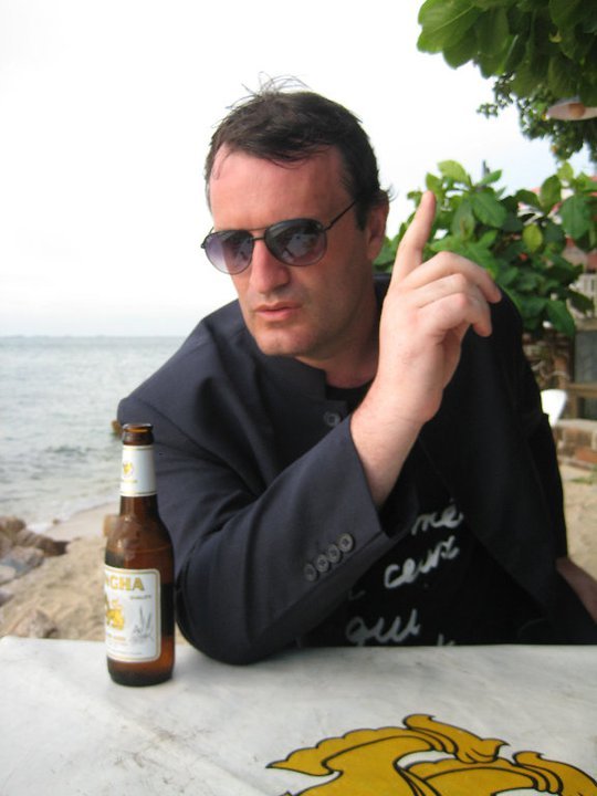 MUFF Festival Director Richard Wolstencroft sitting by the sea, raising a finger while holding a beer on the table.